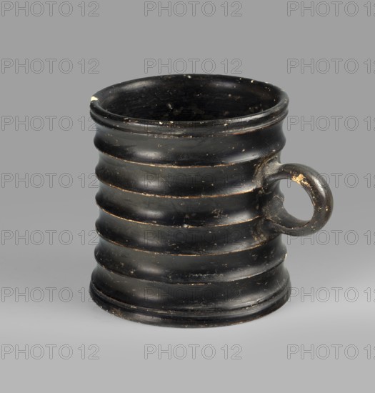 Dice cup. Small cup with a flat slightly concave bottom; walls slightly tapered; distinct rim and foot. Outer surface decorated with seven wide ribs. Entire object covered with black varnish shiny but worn matte in places; small unvarnished fragment on inner surface and outer bottom shows concentric decoration. Traces of turning visible under varnish. Spout slightly chipped; surface shows dirt and secondary losses; varnish worn on rib ridges. Clay color 5 YR 6/6 reddish yellow; varnish color 2.5 YR 2.5/1 black. 5th century BC, Attica. Vessel, cup; height 8.3 cm, width 10.6 cm, depth 8.3 cm., A dark, ribbed cylindrical vessel with a rounded rim and a circular handle on one side, showing worn spots and small chips; the vessel appears black with areas where underlying lighter tones show through, against a neutral gray background.