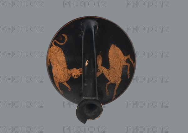 Red-figure askos, Attica, 5th–4th century BC. Vessel askos, height 7.0 cm, width 9.1 cm. Shallow, not very high reservoir with a bow-shaped handle, low cylindrical neck, and spout with outwardly turned edges; flat bottom. Decoration: covered with glossy black varnish; panther facing right with tail curled and paws tucked, and a fawn/goat facing left, running, painted on the shoulders. Thin red line 1–2 cm wide runs around the vessel at the edge of the arms and side walls. Inner rim surface covered with varnish. Technology: thick, high-quality glossy varnish; well-fired; visible traces of turning; cleaned clay free of admixtures. Condition notes: secondary chips on surface; fragment of rim missing; dirty bottom; visible remains of red sealing wax. Color of clay: 5 YR 6/6 reddish yellow. Color of varnish: 10 YR 2/1 black., A round, shallow ceramic object with a single central handle and a short spout is shown from above, featuring a predominantly black surface with worn areas revealing a...