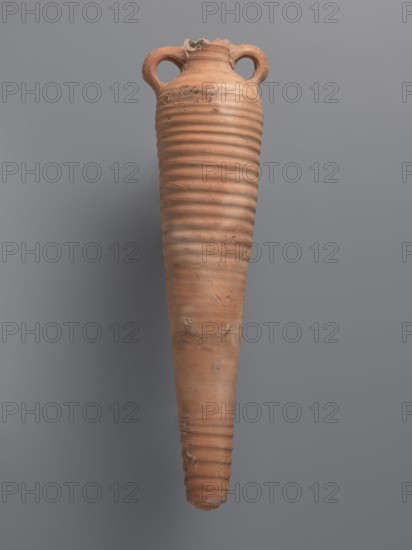 Amphora, unknown Aswan workshop. Spindle-shaped amphora made of pink kaolin clay Gempeler clay IA, 6th–7th century. Thick, ring-shaped vises protruding beyond the vessel outline are attached to the base of the neck and the lower part of the arms. The upper half and lowest section have a fluted surface traces of turning. The bottom is conical, flat-topped. Outer surface smoothed and covered with red engobe, in places burnt to a cream-orange color. Interior coated with black resin impregnation. Material: kaolin clay. Function: storage vessel Coptic amphora. Find location: Edfu. Dimensions: height 50.8+ cm preserved; neck diameter 5.5 cm; body diameter 12.4 cm., A tall, elongated ceramic vessel with a narrow pointed base and a wider rounded top, featuring a ribbed texture along most of its body and a rough, chipped rim with two looped handles near the top; the surface shows patches of wear and uneven texture. The visible colors are terracotta, brown, beige, gray, and muted orange, set aga...