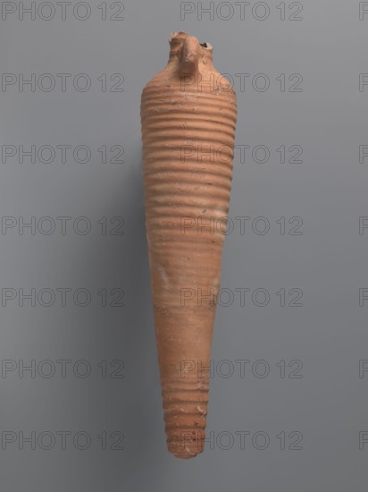 Spindle-shaped amphora, unknown Aswan workshop, 6th–7th century. Thick, ring-shaped vises protruding beyond the vessel outline are attached at the base of the neck and lower part of the handles. The upper half and lowest section have a fluted surface traces of turning. Conical, flat-topped bottom. Outer surface smoothed and covered with red engobe, in places fired to a cream-orange color. Interior coated with black resin impregnation. Made of pink kaolin clay Gempeler clay IA. From Edfu; amphora, height 50.8+ cm preserved, neck diameter 5.5 cm, body diameter 12.4 cm., A tall, narrow terracotta vessel with a ribbed, textured surface and a tapered, pointed bottom suspended against a smooth gray background, showing a broken, irregular rim at the top and an overall weathered appearance with patches of lighter and darker brownish-orange and small areas of pale residue.
