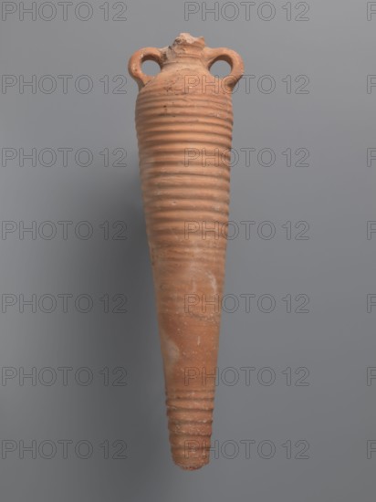 Spindle-shaped amphora, unknown Aswan workshop; 6th–7th century. Kaolin clay pink, Gempeler clay IA. Thick ring-shaped handles protruding beyond the vessel outline are attached at the base of the neck and lower part of the shoulder. Upper half and lowest section have a fluted surface traces of turning. Conical, flat-topped base. Outer surface smoothed and covered with red engobe, in places fired to a cream-orange color; interior coated with black resin impregnation. Function: storage vessel. Preserved height 50.8+ cm; neck diameter 5.5 cm; body diameter 12.4 cm. Find location: Edfu. Style/typology: Aswan/Aswan amphorae, Egyptian/Coptic ceramics., A tall, narrow earthenware vessel with a tapered, elongated body and a rough, ribbed surface, showing a few chips and worn areas; it has a short, irregular neck with a broken rim and a pair of rounded loop handles near the top; the surface is predominantly terracotta orange with some lighter and darker patches and small whitish weathered spots...