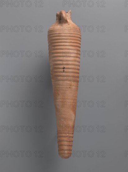 Amphora, unknown Aswan workshop. Spindle-shaped amphora made of pink kaolin clay Gempeler clay IA, 6th–7th century. Thick, ring-shaped vises protrude beyond the outline of the vessel and are attached to the base of the neck and the lower part of the arms. Upper half and lowest section have a fluted surface traces of turning. Conical, flat-topped bottom. Outer surface smoothed and covered with red engobe, in places burnt to a cream-orange color. Interior coated with black resin impregnation. Find location: Edfu. Vessel amphora, height 50.8+ cm preserved, neck diameter 5.5 cm, body diameter 12.4 cm., A tall, tapered clay vessel with a pointed bottom and a narrower top, featuring horizontal ribbed bands running along its length; the surface shows rough texture and small imperfections, with a broken or irregular rim at the top. The visible colors are terracotta, light brown, and hints of darker brown against a neutral gray background.