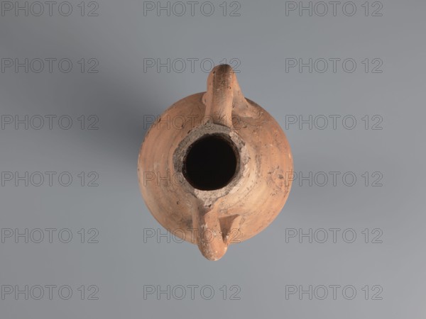 Spindle-shaped amphora, 6th–7th century. Made of pink kaolin clay Gempeler clay IA. Thick, ring-shaped vises protruding beyond the outline are attached to the base of the neck and the lower part of the handles. Upper half and lowest section have a fluted surface traces of turning. Conical, flat-topped bottom. Outer surface smoothed and covered with red engobe, in places burned to a cream-orange color. Interior coated with black resin impregnation. Amphorae type associated with an Aswan workshop; Egyptian/Coptic ceramic storage vessel. Find location: Edfu. Preserved height 50.8+ cm; neck diameter 5.5 cm; body diameter 12.4 cm., A top-down view of a round clay vessel with a central wide opening and a handle-like projection, showing a weathered surface with visible rough textures and chips; the vessel is predominantly terracotta with shades of brown and orange, and the background is a smooth gray.