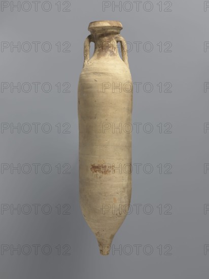 Amphora spatheion, unknown North African workshop Tunisia/Libya. Cylindrical, elongated amphora with a smooth yellow-green surface, probably covered with a coating produced during firing natural engobe, so-called self-slip. Two-stage profiled spout with a rounded top and an outward-sloping lip. Small loop-shaped handles glued to the arms and to the neck just below the spout; conical bottom. Inner walls covered with a resin impregnation, also visible on the outer walls of the upper part of the amphora. Complete vessel, ceramic body not visible. 2nd half of the 6th–7th century; findspot: Edfu. Vessel amphora. Height 44.2 cm; diameter 6.6 cm rim, 10 cm body., A tall, elongated ceramic vessel with a narrow neck and a small rounded foot, showing a pair of looped handles near the top; the surface is mostly beige with areas of darker brown and light tan, and there are a few small reddish-brown spots and subtle variations in texture on the body, set against a smooth gray background.