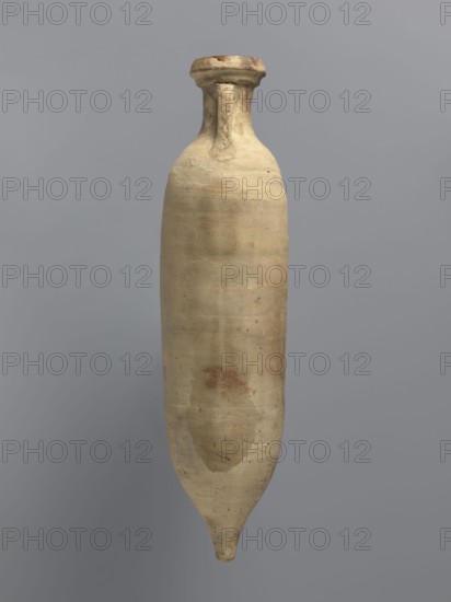 Amphora spatheion, unknown North African workshop Tunisia/Libya. Cylindrical, elongated amphora with a smooth yellow-green surface, probably covered with a coating produced during firing natural engobe, self-slip. Two-stage profiled spout with a rounded top and an outward-sloping lip. Small loop-shaped handles attached to the arms and to the neck just below the spout; conical bottom. Inner walls covered with a resin impregnation, also visible on the outer walls of the upper part of the amphora. Complete vessel, ceramic body not visible. Date: 2nd half of the 6th–7th century. Place of discovery: Edfu. Function/type: vessel, amphora spatheion. Dimensions: height 44.2 cm; rim diameter 6.6 cm; body diameter 10 cm., A tall, narrow ceramic vessel with a rounded body tapering to a pointed base is shown against a plain background; the vessel has a short neck with a flattened, flared rim and visible surface irregularities and worn spots, with overall beige and tan tones and a few areas of darke...