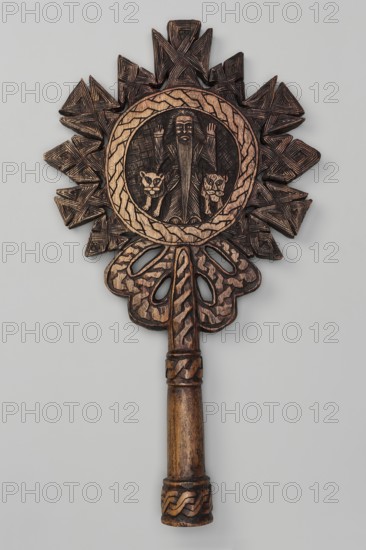 Processional cross, Ethiopian workshop. In the center of the cross, in the tondo, a standing man is depicted frontally with hands raised in an orant gesture. On his sides stand a lion and a lioness. The man has a long, ankle-length beard—likely the hermit Paulos. The tondo is bordered by a wide braid-decorated band from which radiate small crosses with rhomboid arms. The short, sleeve-shaped handle is decorated with two rings with a braided ornament. The reverse shows a winged figure above two birds. 20th century, Ethiopia. Cross; height 44 cm, width 24 cm., A carved wooden object with a long cylindrical handle and an ornate top featuring a circular medallion surrounded by a radiating geometric design; the carving includes a central figure with two animal faces flanking it, all rendered in carved texture and shallow relief. The surface shows visible carved lines and crosshatching, braided motif bands around parts of the handle, and carved openwork shapes beneath the medallion. The colo...
