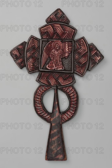 Processional cross, Ethiopian workshop. At the intersection of the cross's arms is a square depicting three seated, frontally posed figures of the Holy Trinity, each holding a spherical object in their right hand as a symbol of power over the world. The arms of the cross are trapezoidal with rounded outer sides; the horizontal beam terminates in trapezoidal crosses, and a similar cross is placed at the top of the vertical beam. The handle is sleeve-shaped, widening at the bottom, with two semicircles attached to its sides. On the reverse, at the intersection of the arms, there is an archangel holding a sword. 20th century, Ethiopia. Material: metal cross. Height 39 cm, width 23 cm., An intricately carved wooden object with a cross-like top section and a long tapered handle, featuring a central square panel containing a carved human figure with wings; the surface shows carved geometric patterns and textured areas, with visible colors including dark brown, reddish-brown, black, and a pal...