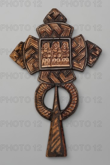Processional cross, Ethiopian workshop. At the intersection of the cross's arms is a square depicting three seated, frontal figures of the Holy Trinity, each holding a spherical object in their right hand as a symbol of power over the world. The arms of the cross are trapezoidal with rounded outer sides; the horizontal beam terminates in trapezoidal crosses. A similar cross is placed at the top of the vertical beam. The handle is sleeve-shaped, widening at the bottom, with two semicircles attached to its sides. On the reverse, at the intersection of the arms, there is an archangel holding a sword. 20th century, Ethiopia. Cross; height 39 cm, width 23 cm., A carved wooden object with a vertical handle and a circular loop near its top features a cross-shaped upper section with a rectangular central panel showing a group of closely spaced seated figures; the surface displays carved linear and geometric patterns with a textured, slightly rough appearance and visible wear. The colors visibl...