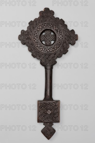 Hand-made equal-armed cross inscribed in a circular frame, surrounded by a braided ornament arranged in a rhomboid shape. The space between the arms of the cross and the frame is cut out. The oval, smooth handle merges into a rectangular plaque topped with a rhomboid element. The plaque and rhomboid element are decorated with braid. Hand-made Ethiopian cross, 20th century, Addis Ababa. Height 44 cm, width 20 cm., A carved wooden object with an elongated handle and an ornate top section, predominantly dark brown with subtle lighter brown highlights, featuring a circular open center surrounded by geometric and interlaced carved patterns, a square carved element near the lower end with braided motifs, and a small carved diamond-shaped extension at the very bottom, all set against a light gray background.