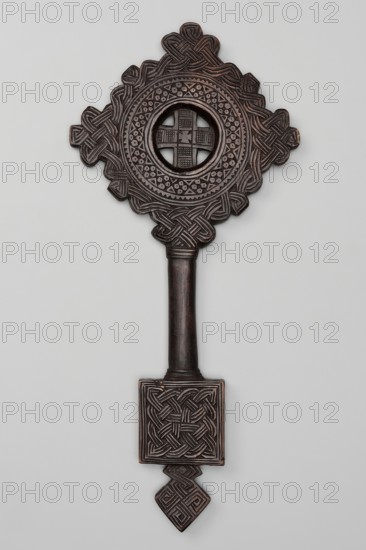 Hand-made cross, Ethiopian workshop. The equal-armed cross is inscribed in a circular frame, surrounded by a braided ornament arranged in a rhomboid shape. The space between the arms of the cross and the frame is cut out. The oval, smooth handle merges into a rectangular plaque, topped with a rhomboid element. The plaque and the rhomboid element are decorated with braid. 20th century, Addis Ababa. Cross; height 44 cm, width 20 cm., A carved wooden object in dark brown tones with a long handle and an ornate top, featuring interlacing knot patterns and geometric motifs; the top has a circular open center surrounded by concentric decorative bands and a quatrefoil-like outer shape, and the lower end transitions into a square panel with braided carving and a small diamond-shaped finial, all set against a pale gray background.