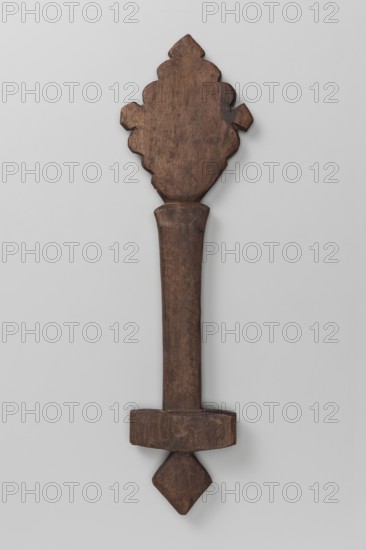 Hand-made cross, Ethiopian workshop. Hand-made cross — very schematic. It consists of a rhombus mounted on a shaft topped with a rectangular plate with a rhomboid projection growing from the center of the plate's lower side. The upper, rhombus-shaped part is decorated with wavy incisions on the edges and rhomboids on the tops. 20th century, Tigray. Cross; height 29 cm, width 8 cm., A carved wooden object with a dark brown surface against a light gray background, showing a broad ornate top with scalloped edges, a long tapered central shaft with faint carved patterns, a rectangular crosspiece near the lower end, and a diamond-shaped finial at the bottom; the wood displays varied brown tones and visible grain and worn areas.