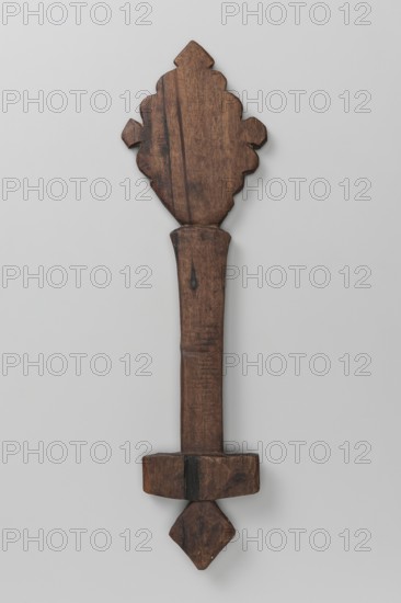 Hand-made Ethiopian cross, 20th century. Very schematic design: a rhombus mounted on a shaft topped with a rectangular plate with a rhomboid projection growing from the center of the plate's lower side. Upper rhombus-shaped part decorated with wavy incisions on the edges and rhomboids on the tops. Material: hand-made; place of manufacture: Tigray. Cross; height 29 cm, width 8 cm., A wooden object with a carved decorative top, a long rectangular shaft, and a geometric lower end, all shown against a neutral pale gray background; the wood displays various brown tones with darker brown grain and spots, and there are subtle variations in the brown surface suggesting wear and texture.