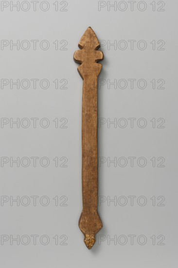 Hand cross, Ethiopian workshop. A small, roughly detailed hand cross with a rhomboid ending—shaped like a spear. Notches on the rhomboid suggest the crossbar and vertical beam. The long shaft ends in a rhomboid at the bottom. 20th century, Tigray. Cross; height 34 cm, width 4.2 cm., A long, narrow wooden object with a carved decorative top and bottom, showing a warm brown wood grain and a slightly darker brown patina, set against a pale gray background.