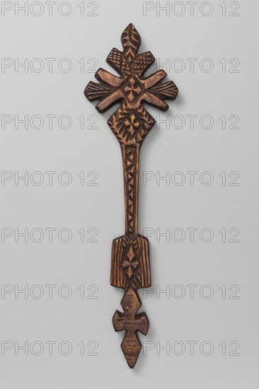 Hand cross, Ethiopian workshop. The upper part, inscribed in a rhombus, is mounted on a long shaft terminating at the bottom with a rectangular plaque; below it is decorated with a cross with rhomboid arms. The cross is covered with dense geometric ornament; in the central part is a small cross rosette. The side and upper arms are stylized as flower petals a schematic lily. The lower arm is rhombus-shaped with cross rosettes. The shaft is decorated with incisions in a broken-line pattern. The plaque has a cross rosette in the middle and incisions parallel to its longer edges. 20th century, Addis Ababa. Cross; height 36.5 cm, width 9.5 cm., A carved wooden object with a long narrow handle and decorative ends, displaying a rich brown tone with darker brown carved recesses and lighter brown highlights; the top end fans into a symmetrical, leaf-like cluster with carved geometric and linear patterns, while the handle features a vertical series of carved shapes and textured lines, and the bo...