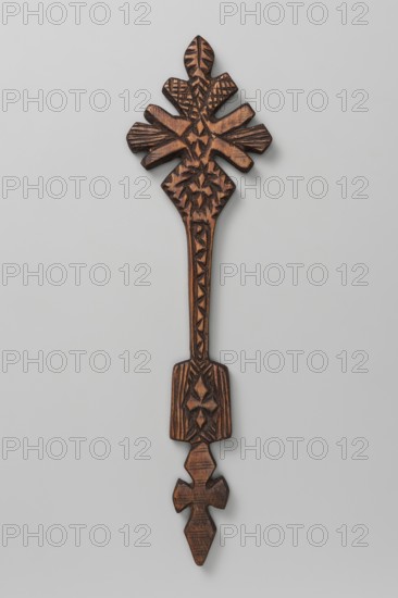 Hand cross, Ethiopian workshop. Hand cross with the upper part inscribed in a rhombus mounted on a long shaft terminating at the bottom with a rectangular plaque; below it is decorated with a cross with rhomboid arms. The cross is covered with dense geometric ornament; in the central part is a small cross rosette. The side and upper arms are stylized as flower petals schematic lily. The lower arm is rhombus-shaped with cross rosettes. The shaft is decorated with incisions forming a broken line. The tablet has a cross rosette in the middle and incisions parallel to its longer edges. 20th century, Addis Ababa. Cross; height 36.5 cm, width 9.5 cm., A carved wooden object with a vertical elongated shape featuring decorative carved patterns and geometric motifs, shown against a light gray background; the wood displays shades of brown with darker brown carved lines and highlights, and there are a few symmetrical protruding elements near the top and bottom, with carved textures and triangular...