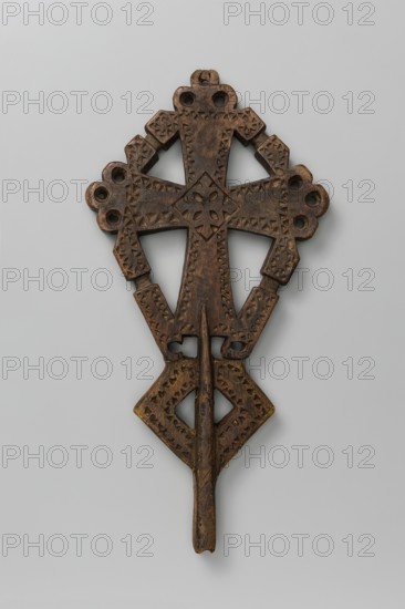 Processional cross, Ethiopian workshop. A small, likely processional isosceles cross with slightly widening arms terminating in trefoil-shaped projections except for the lower arm, which has a mounting socket. Arms are connected by crossbars with a central recess on each. Surface decorated with incised small triangles forming an edge ornament. At the arms' intersection is a rhombus containing a rosette and central cross; lower part features a rhombus through which the socket runs, its edges likewise decorated with triangular incisions. 20th century, Addis Ababa. Height 29 cm, width 14.5 cm., A carved wooden object with a symmetrical, cross-like shape featuring geometric cutouts and incised triangular patterns, displaying a rich brown color with areas of darker brown and hints of yellowish tones.