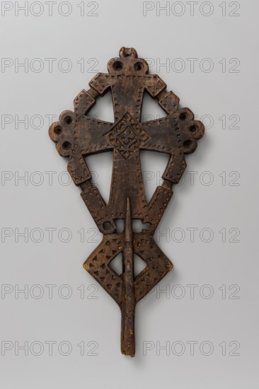 Processional cross, Ethiopian workshop. A small isosceles processional cross with slightly widening arms that terminate in trefoil-shaped projections except the lower arm, which has a narrow socket for mounting. The arms are connected by crossbars, each with a crossbar recess at its center. Surface decorated with incised small triangles forming an edge ornament. At the intersection of the arms is a rhombus containing a rosette and central cross; below is a rhombus pierced by the socket. 20th century. Made in Addis Ababa. Cross; height 29 cm, width 14.5 cm., A carved wooden object with a dark brown surface and lighter brown highlights, shaped like a stylized cross with a long tapered stem; the top section has rounded lobes and circular cutouts, the arms of the cross are broad with triangular notches along their edges, and a diamond-shaped raised motif sits at the center; a geometric openwork pattern appears in the lower section, and the piece shows carved triangular punctures and incise...