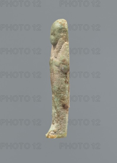 Ushabti, Mummy-shaped figurine, funerary gift, hoes. Hands folded on the chest, uncrossed, right hand under left; both hands hold hoes. Smooth tripartite wig with a headband. Facial details poorly legible except for large eyes. Smooth back. No inscription. c. 1069–c. 656 BC, Third Intermediate Period, Egypt. Height 6.4 cm., A slender vertical figurine-like object with a rounded top and a narrow base, standing against a plain gray background; the surface shows a rough, weathered texture with patches of pale green, light brown, and beige, along with some darker brown spots and small areas of lighter yellow near the base.