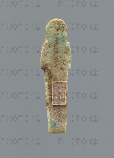 Ushabti, Mummy-shaped figurine, funerary gift, funerary hoe. Hands folded on the chest, uncrossed, right hand under left; both hands hold hoes. Smooth tripartite wig with a headband; facial details poorly legible except for large eyes. Smooth back. No inscription. c. 1069–c. 656 BC Third Intermediate Period, Egypt. Height 6.4 cm., A worn, elongated figurine-like object stands vertically against a plain gray background, showing a rounded head, narrow shoulders, and a tapering body; the surface is rough and pitted with an aged texture, and a small rectangular fabric patch is affixed to the lower torso area. The visible colors include muted green, pale blue, tan, brown, and hints of reddish discoloration, with darker brown spots and lighter sandy areas across the form.