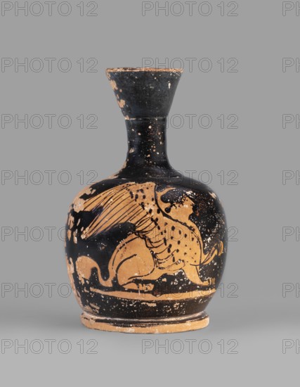 Lekythos with a sphinx. A small Attic red-figure lekythos with a viscous vise? may be mistranscribed. The vessel has a narrow neck, a rounded belly mounted on a round foot. The central part of the front belly is decorated with an image of a sphinx; the back of the vessel has worn finials. 1st half of the 4th century BC, Attica. Ceramic vessel, height 9.4 cm, diameter 6 cm., A rounded ceramic vessel with a narrow neck and flared rim sits against a plain background. The vessel shows a worn black surface with areas where a tan clay color is exposed, forming a large painted figure wrapping around the body. The figure appears to have a curved body, a long tail, and a wing-like element rendered with fine black lines on the tan ground. The base and rim show chipped tan edges where the black slip has worn away. The overall visible colors are black and tan with spots of lighter wear.