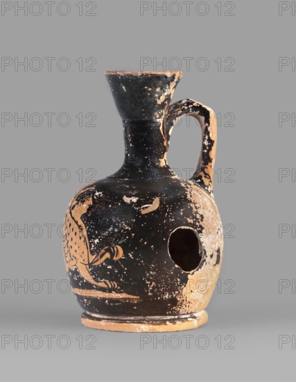 Lekythos with a sphinx, unknown Attic workshop. A small Attic red-figure lekythos with a spout. The neck is narrow; the belly is round, mounted on a round foot. The central part of the front of the belly is decorated with an image of a sphinx; the back has worn finials. 1st half of the 4th century BC, Attica. Vessel; height 9.4 cm, diameter 6 cm., A rounded ceramic vessel with a narrow neck, flared rim, and a single handle connecting rim to shoulder sits against a plain gray background; the surface shows a worn black slip with areas of exposed terracotta creating a mottled pattern, and there is a large round hole on one side of the body; traces of a painted animal motif are visible where the black slip remains, and the handle and rim display chipped edges and flaked areas revealing the underlying tan color.