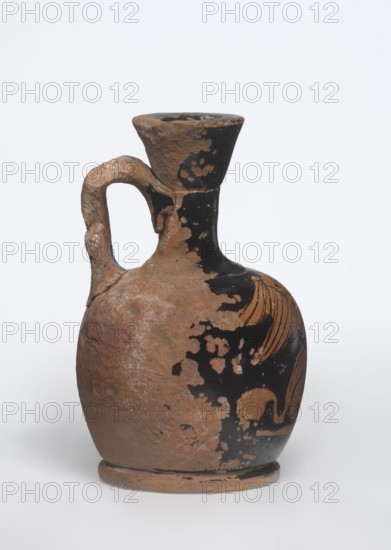 Lekythos with a sphinx. Small Attic red-figure lekythos with a vise. Narrow neck, round belly on a round foot. Central front of the belly decorated with an image of a sphinx; back of the vessel has worn finials. 1st half of the 4th century BC, Attica. Ceramic vessel Greek, height 9.4 cm, diameter 6 cm. Maker: unknown Attic workshop., A worn ceramic jug with a rounded body, narrow neck, flared rim, and a curved handle attached from neck to shoulder; the surface shows patches of black slip partly flaked away to reveal an underlying reddish-brown clay, with additional areas of lighter brown and pale weathered spots; a portion of the black decoration forms curved lines on one side, and the jug sits on a simple circular foot on a neutral light background.