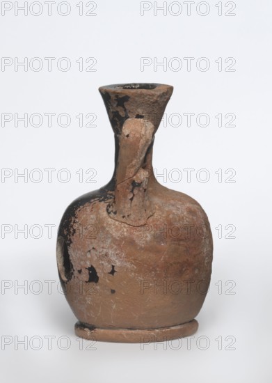Lekythos with a sphinx, unknown Attic workshop. A small Attic red-figure lekythos with a vise. The neck of the vessel is narrow. The belly is round, mounted on a round foot. The central part of the front side of the belly is decorated with an image of a sphinx. The back of the vessel has worn finials. 1st half of the 4th century BC, Attica. Vessel; height 9.4 cm, diameter 6 cm. Materials: ceramics; subject/iconography: sphinx., A worn ceramic vessel with a rounded body, narrow neck, and flared rim, showing a rough, pitted surface and irregular patches where a dark coating has peeled away; the form includes a single handle attaching the neck to the shoulder and a raised circular base. The visible colors are earthy brown, dark black, and lighter beige-gray areas where the surface is abraded.