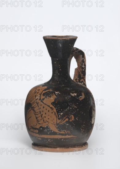 Lekythos with a sphinx, unknown Attic workshop. A small Attic red-figure lekythos with a vise. The neck of the vessel is narrow. The belly is round, mounted on a round foot. The central part of the front side of the belly is decorated with an image of a sphinx. The back of the vessel has worn finials. 1st half of the 4th century BC, Attica. Vessel; ceramics; Greek ceramics; sphinx mythological/iconographic. Height 9.4 cm, diameter 6 cm., A small rounded ceramic vessel with a narrow neck, flared rim, and single curved handle, showing areas of wear and chipped edges; its surface features a painted animal motif with the body and legs rendered in a tan color against a black background, with speckled and worn black and tan patches across the vessel, and a pale rim and base edge revealing the underlying clay color.
