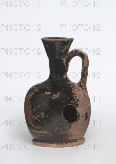 Lekythos with a sphinx. Attic red-figure lekythos, 1st half of the 4th century BC, Attica. Small ceramic vessel with a narrow neck, round belly on a rounded foot; central front of the belly decorated with an image of a sphinx; back of the vessel has worn finials. Height 9.4 cm, diameter 6 cm. Maker unknown Attic workshop., A small ceramic jug with a rounded body, a narrow neck that flares into a wide rim, and a single curved handle; the surface shows worn areas and chipped paint, revealing a reddish-brown clay beneath a predominantly black slip, with patches of the reddish-brown visible across the body and handle, and a circular hole on one side; there is a painted decorative element in a reddish-brown hue against the black on the body, and the base shows exposed reddish-brown clay with some black residue.