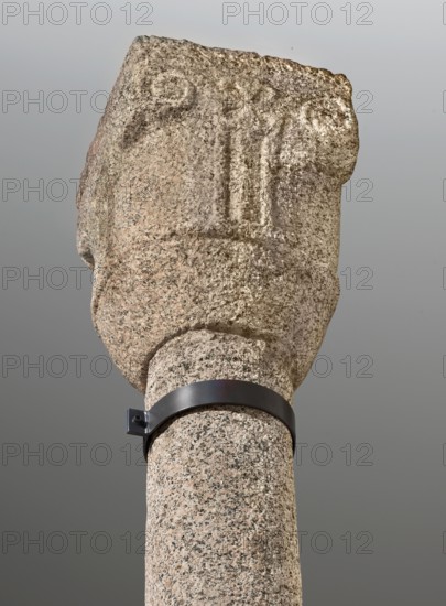 Column, Nubian workshop; architectural element with relief decorations and crosses; monolithic base cuboid 57 x 51 cm topped by a truncated cone 96 cm high; cylindrical monolithic shaft slightly narrowed at top with a wide slightly convex half-cylinder encircling base and two half-cylinders below the capital; low relief at two-thirds height depicting a cross with equal, widening triangular arms inscribed in a circle; capital 72 cm high with cup-shaped lower part, then cylindrical section transforming into a quadrangle covered with a rectangular bakusa plate 57 x 52 cm; corners of the echinus decorated with four volutes forming protruding corners and, between the volutes on one side, a low relief depicting a cross with equal triangular arms inscribed in a circle; 2nd half of the 7th–1st quarter of the 8th century; Faras Sudan; height 492 cm; diameter 51 cm., A close-up of a tall stone column topped with a roughly carved stone head, showing a textured surface with speckled tones of beige...
