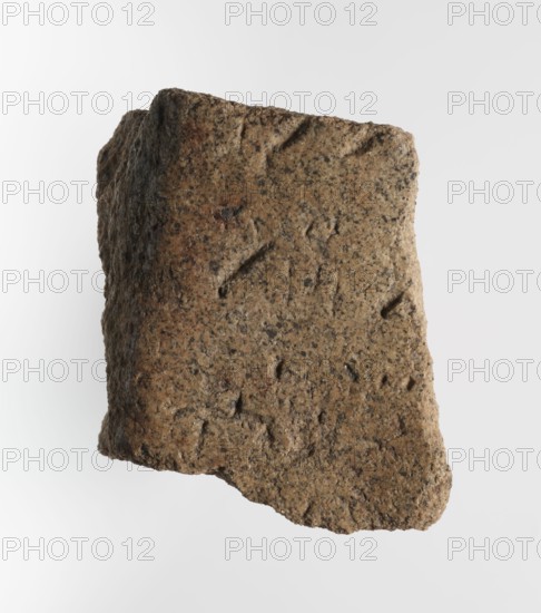 Inscription - fragment, unknown,  craftsman, inscriptions, Granite fragment with engraved hieroglyphic inscription, 760–653 BC; Third Intermediate Period; 25th Dynasty, Old Dongola type of site / place of finding, inscription, height 8 cm, width 7 cm, A roughly rectangular piece of rough, porous material with an uneven, jagged edge, shown against a plain light background. The surface is textured with small pits and irregularities and shows a grainy, speckled appearance. The visible colors include shades of brown, tan, beige, and darker flecks of black or dark gray, with subtle warmer brown patches in places. The object has areas of shadow along one side indicating depth and uneven thickness, and the top edge appears slightly rounded while the bottom edge tapers irregularly.