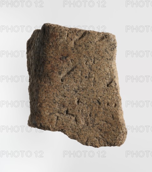 Inscription - fragment, unknown,  craftsman; inscriptions. Granite fragment with engraved hieroglyphic inscription, 760–653 BC; Third Intermediate Period; 25th Dynasty. Found at Old Dongola. Inscription. Height 8 cm, width 7 cm., A rough, irregularly shaped stone fragment floats against a light gray background, showing a coarse, pitted surface with an uneven edge. The stone displays a mix of brown and tan tones with darker brown and black speckling throughout, and small depressions and ridges create a textured appearance. The fragment has a slightly curved profile with visible granular texture and subtle variations in color intensity across its face, including some warmer brown patches and cooler grayish areas.