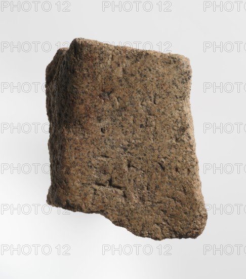 Inscription - fragment, unknown,  craftsman; inscriptions; granite fragment with engraved hieroglyphic inscription; 760–653 BC; Third Intermediate Period; 25th Dynasty; Old Dongola type of site / place of finding; inscription; height 8 cm; width 7 cm, A rough, irregularly shaped piece of rock or stone with a textured surface is shown against a light background. The stone displays a mix of brown and tan tones with speckles and patches of darker gray and black. Its surface has small pits, grooves, and uneven edges, and a few areas show subtle reddish-brown discoloration. The overall form is blocky but asymmetrical, with one edge appearing more angular and the opposite side more rounded.