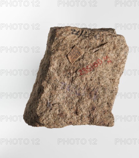 Inscription - fragment, unknown,  craftsman; inscriptions. Granite fragment with engraved hieroglyphic inscription. 760–653 BC; Third Intermediate Period; 25th Dynasty. Old Dongola type of site / place of finding. Inscription. Height 8 cm, width 7 cm., A rough, irregularly shaped rock fragment occupies the image, showing a coarse, granular surface texture with visible grains and small pits. The rock displays a mix of colors including tan, beige, light brown, and speckles of darker brown and black, with subtle hints of gray and faint areas of purple. A small square patch of a different, slightly darker brown material is attached near the upper central area. The background is a smooth gradient of white to very light gray.