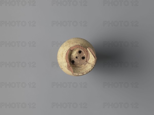 Water vessel qulla. Ovoid, elongated belly with the edge of the foot protruding outward. Short, wide neck transitions into a higher, slightly flared section above a sharp break in the wall. Inside the neck is a strainer formed by puncturing holes in the clay before firing. Entire surface covered with cream-yellow whitewash. Made of medium-porous Nile clay. 2nd half of the 5th–6th century. From Tell Atrib. Vessel for storing water. Height 17.2 cm; width 9.3 cm; outer rim diameter 7 cm; base diameter 5 cm., A single round ceramic object sits near the center of the image against a smooth flat background; the object is mainly beige with areas of reddish-brown where the surface is chipped or worn, and it has a rough, speckled texture. The top face of the object shows a recessed circular area with several circular holes and fragmented edges, and a shadow extends to the right creating a gradient from medium gray near the object to darker gray farther out. The background is uniformly gray with...
