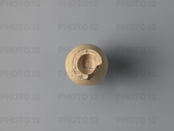 Water vessel qulla. Ovoid with elongated belly; edge of foot protrudes outward. Short, wide neck leading to a higher, slightly flared section above a sharp break in the wall. Strainer inside the neck formed by punctured holes in the clay before firing. Entire surface covered with cream-yellow whitewash. Medium-porous Nile clay. 2nd half of the 5th–6th century. Vessel for storing water. Height 17.2 cm; width 9.3 cm; outer rim diameter 7 cm; base diameter 5 cm. Egyptian workshop; Tell Atrib., A round, broken pottery object viewed from above sits near the center of a smooth grey surface, showing textured beige and light brown clay with rough edges and a missing chunk along one side, casting a soft shadow to the right; visible colors include beige, light brown, pale tan, grey, and darker grey in the shadow.