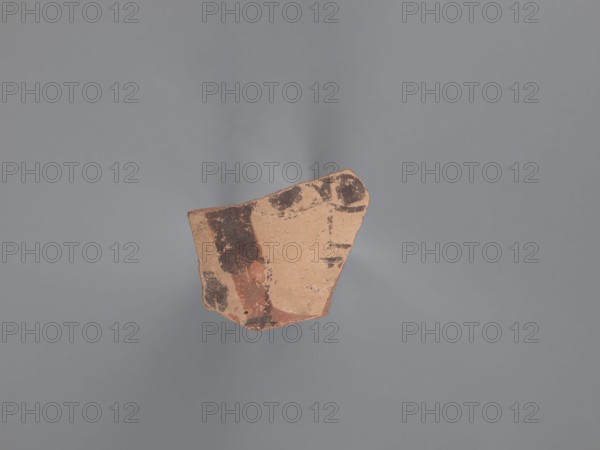 Fragment of the body of a vessel with decoration painted in black and red against a pink-beige engobe background. Around the face, preserved only on the right side of the head, is wavy black hair covering the ear. The black pupils are surrounded by an almond-shaped outline resembling the shape of the eye. The nose is represented by a long vertical and a short horizontal line; the mouth by two unequal lines. A round black dot is next to the face. Nile clay of medium porosity. 5th–6th century, Egyptian workshop, Tell Atrib, painted Coptic vessel with figural decorations; height 3.2 cm, width 4 cm., A fragment of pottery with a rough, irregular shape sits against a plain gray background. The pottery piece is mainly a tan or light brown color with areas of darker brown and reddish-brown pigment forming irregular patches and worn shapes. The surface shows texture variations and some chipped edges, and the lighting creates soft shadows around the fragment.