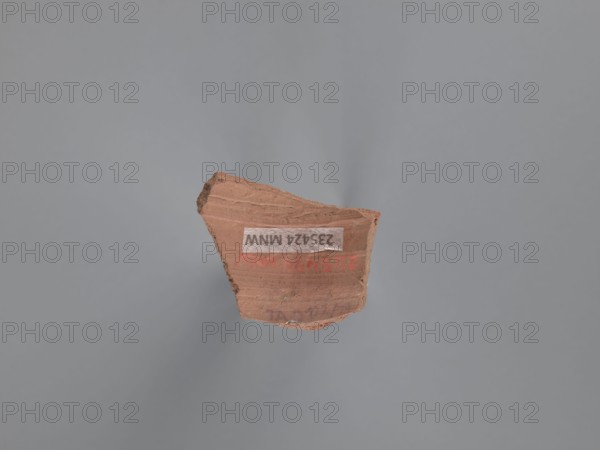 Fragment of a vessel, painted Coptic vessel with figural decoration, Fragment of the body of a vessel with decoration painted in black and red against a pink-beige engobe background. Around the face, preserved only on the right side of the head, is wavy black hair covering the ear. The black pupils are surrounded by an almond-shaped outline resembling the shape of the eye. The nose is represented by a long vertical and a short horizontal line; the mouth by two unequal lines. A round black dot is next to the face. Nile clay of medium porosity. 5th–6th century, Tell Atrib, vessel, height 3.2 cm, width 4 cm, A fragment of reddish-brown pottery with rough, uneven edges sits against a smooth gray background; the pottery piece shows layered texture and slight ridges on its surface and has areas of darker brown along one edge, while small patches of lighter tan appear where the surface is worn.