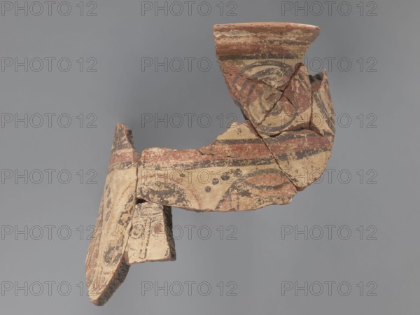 Painted storage vessel, fragment of the upper part of a large storage vessel: the neck and upper part of the body. Decoration painted in black and red on a creamy white underlay. Rim has a thick, outward-protruding edge with a concave outer surface. Upper part painted with a row of horizontally running spirals with large red spots; spirals enclosed in red horizontal stripes, each bounded by black lines. Below, partially on the body, are figural representations: heads or fragments of male or female figures; the central head is surrounded by a halo. Space between the heads is filled with floral elements in the lower part and with black dots in the upper part. The heads on the sides wear headgear and have no halos. Made of Nile clay, medium porous N.B2. 6th century, Egypt. Vessel; diameter 26 cm external rim, height 31 cm., A broken ceramic fragment with curved and irregular edges suspended against a gray background, showing painted geometric and circular patterns in shades of red, beige,...