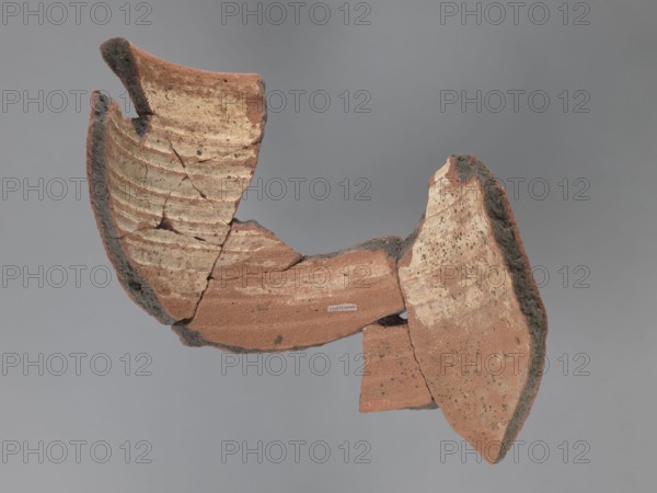 Painted storage vessel, painted Coptic storage vessel with figural and floral decorations. Fragment of the upper part of a large storage vessel: the neck and upper part of the body. Decoration painted in black and red on a creamy white underlay. The rim has a thick, outward-protruding edge with a concave outer surface. Upper part painted with a row of horizontally running spirals with large red spots; spirals enclosed in red horizontal stripes, each bounded top and bottom by black lines. Below, partially on the body, are figural representations: heads or fragments of male and female figures. The central head is surrounded by a halo; the side heads wear headgear and have no haloes. Spaces between heads filled with floral elements in the lower part and black dots in the upper part. Vessel made of Nile clay, medium porous N.B2. 6th century, Egypt; vessel; diameter 26 cm external rim; height 31 cm., A broken ceramic object made of reddish-brown clay with rough dark gray edges is shown agai...