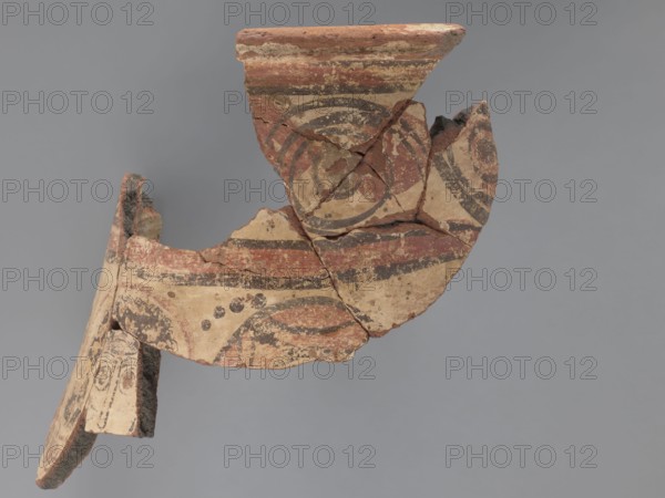 Painted storage vessel, storage vessels, painted Coptic vessels, figural decorations, floral decorations. Fragment of the upper part of a large storage vessel: the neck and upper part of the body. The decoration is painted in black and red on a creamy white underlay. The rim of the vessel has a thick, outward-protruding edge with a concave outer surface. The upper part of the vessel is painted with a row of horizontally running spirals, on which large red spots are painted. The spirals are enclosed in red horizontal stripes, each bounded at the top and bottom by black lines. Below, partially on the body, are figural representations: heads, or fragments of male or female figures. The central head is surrounded by a halo. The space between the heads is filled with floral elements in the lower part and with black dots in the upper part. The heads on the sides are wearing headgear - there are no haloes. Vessel made of Nile clay, medium porous N.B2. 6th century, Egypt, vessel, diameter 26 c...