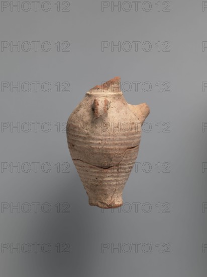 Water vessel qulla, bottle, Coptic vessel. Water vessel bottle, gulla. Ovoid, elongated body, wider in the uppermost part, above half the vessel's height. Low foot, hollow inside, poorly defined externally. Two small vises are glued to the arms, and between them, on an axis perpendicular to their attachment, a conical spout for drinking water is glued. The neck, preserved in its lowest part, is separated from the body by a small, sharply profiled hump. The entire surface of the vessel is covered with light whitewash. Nile clay, medium-porous, 5th century, Tell Atrib, vessel, height 17.0 cm, width 11.5 cm, diameter 4 cm base, An aged, weathered clay vessel in shades of beige, tan, and reddish-brown floats against a smooth gray background; the vessel has a rounded, tapering body with horizontal ridged texture, a small protruding spout on one side, an irregular broken rim with rough edges, visible cracks and chips, and surface spots of darker brown and lighter beige variations.