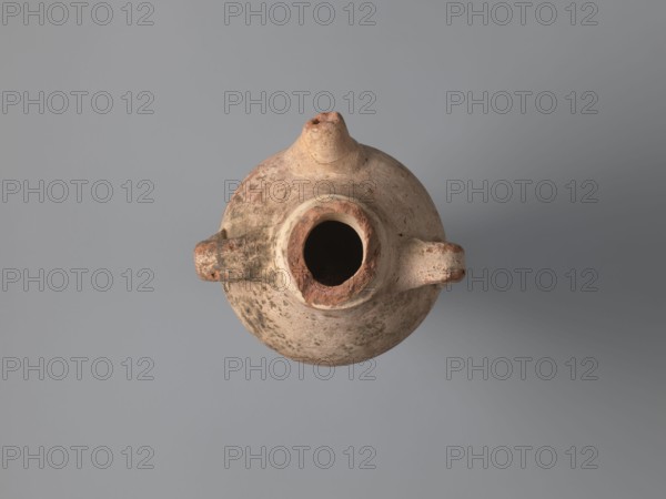 Water vessel qulla, bottles, Coptic vessels, water vessels qullae, qulla, Water vessel bottle, gulla. Ovoid, elongated body, wider in the uppermost part, above half the vessel's height. Low foot, hollow inside, poorly defined externally. Two small vises are glued to the arms, and between them, on an axis perpendicular to their attachment, a conical spout for drinking water is glued. The neck, preserved in its lowest part, is separated from the body by a small, sharply profiled hump. The entire surface of the vessel is covered with light whitewash. Nile clay, medium-porous, 5th century, Tell Atrib, vessel, height 17.0 cm, width 11.5 cm, diameter 4 cm base, A worn round clay vessel viewed from above with a central circular opening and a short tapered spout, two protruding loop handles on opposite sides, a rough textured surface showing patches of darker and lighter areas, and visible surface irregularities; colors include beige, tan, light brown, reddish-brown, and some darker grayish sp...