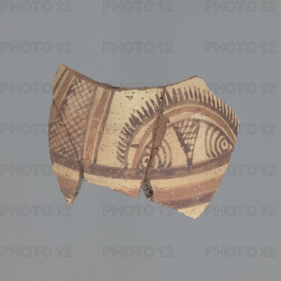 Bottle fragment, unknown Egyptian workshop. Fragment of a small water bottle, probably qulla type. Made of Nile clay type B with significant limestone inclusions and traces of chaff. Fracture nearly uniform dark grey fading to dark grey-brown, 10YR4/1–10YR3/2. Inner surface unworked dark grey, 10YR4/1; outer surface covered with whitewash very pale brown, 10YR8/3 bearing painted brown-red decoration: horizontal stripes, semicircles, lines, concentric circles, grid. 5th–7th century, Egypt. Vessel: height 6.2 cm, width 9.3 cm, thickness 6 mm., A broken ceramic fragment with curved edges and an irregular shape, showing a painted geometric and linear pattern in shades of brown on a beige base; the decoration includes dark brown arcs, concentric curved lines, triangular filled areas, and a banded border, with the surface showing rough, chipped edges and small areas of darker brown where the paint is thicker, all set against a solid gray background.