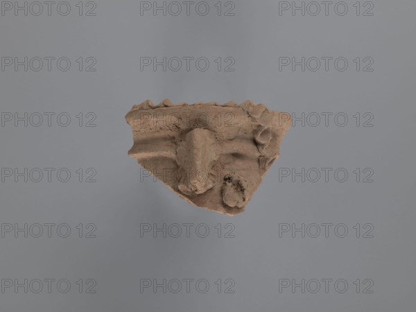 Fragment of a vessel with applied decoration;, Coptic pottery, plastic decoration. Fragment of the neck of a vessel with plastic decoration: the spout has a rounded edge, slightly concave below the bulge. The hand-applied plastic decoration conveys a human face or animal head. A narrow, slightly humped nose is clearly visible and may also have served as a handle. The concave clay bump inside is probably an eye, although it is glued to the neck of the vessel at nostril level. A strip of clay with a series of finger-made indentations imitation of hair? is glued to the top of the spout, descending towards the body. Ceramic body made of Nile clay NA, low porosity. Unfinished surface. 2nd half of the 5th–6th century, Tell Atrib; vessel. Height 7.6 cm, width 12 cm, diameter 16 cm external spout., A fragment of a rough, brown clay object with an irregular triangular shape floats against a smooth gray background, showing textured, uneven surfaces with ridges and folds, a prominent central prot...
