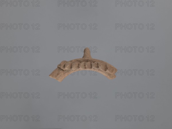 Fragment of a vessel with applied decoration, Coptic pottery, plastic decoration. Fragment of the neck of a vessel with plastic decoration. The spout has a rounded edge, slightly concave below the bulge. The hand-applied plastic decoration conveys features of a human face or animal head: a narrow, slightly humped nose possibly served as a handle and a concave clay bump inside that is probably an eye, glued to the neck at nostril level. A strip of clay with a series of finger-made indentations imitation of hair? is glued to the top of the spout, descending towards the body. Ceramic body made of Nile clay NA, low porosity. Unfinished surface. Dated to the 2nd half of the 5th–6th century, from Tell Atrib; vessel. Height 7.6 cm, width 12 cm, diameter 16 cm external spout., A curved brown clay fragment with a raised central knob and a series of rounded protrusions along its top edge is shown against a smooth gray background; the clay piece has textured, rough surfaces with small chips and d...