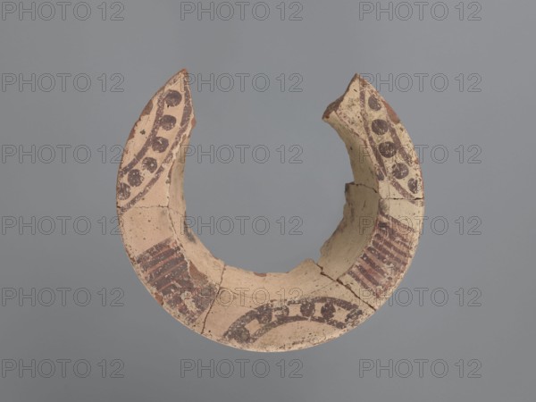 Fragment of a censer, upper part with a spout. The inner and outer surfaces are covered with a creamy whitewash. The spout features two motifs, each repeated three times: 1 a series of 7–9 lines painted on a red background perpendicular to the spout edge; 2 a pair of lines arching against the outer edge of the spout with 5–6 dots between them. Flat top rim with a slightly sloping surface. Pottery body made of Nile clay N.B1, medium porous. Coptic pottery, painted Coptic vessel, 6th–7th century, height 6.7 cm, width 19.2 cm, diameter 19 cm external rim., A broken crescent-shaped ceramic fragment with a pale beige surface and darker brown painted decorative bands, showing a few dotted and striped patterns around the curve; the object has rough broken edges and visible cracks, with areas of flaking and worn paint, set against a uniform gray background.