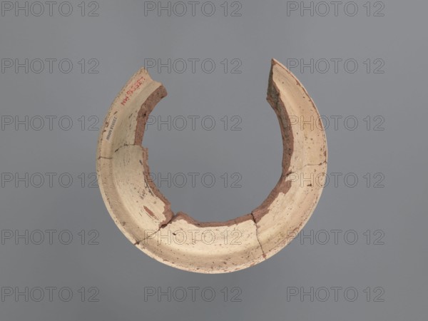 Fragment of a censer, Upper part of the censer with a spout. The inner and outer surfaces are covered with a creamy whitewash. The spout features two types of motifs, each repeated three times: 1 a series of 7–9 lines painted on a red background perpendicular to the edge of the spout; 2 a pair of lines arching against the outer edge of the spout with 5–6 dots between them. Flat top rim with a slightly sloping surface. Pottery body made of Nile clay N.B1, medium porous, 6th–7th century, Tell Atrib, vessel; height 6.7 cm, width 19.2 cm, diameter 19 cm external rim., A broken circular ceramic fragment with rough, uneven edges and visible cracks, showing inner and outer curved surfaces; the fragment has areas of light beige and off-white with patches of reddish-brown clay exposed along the broken edges and inner sections; the background is a uniform muted gray.
