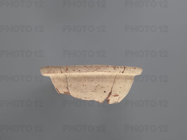 Fragment of a censer, Upper part of the censer with a spout. The inner and outer surfaces are covered with a creamy whitewash. The spout features two types of motifs, each repeated three times: 1 a series of 7–9 lines painted on a red background perpendicular to the edge of the spout; 2 a pair of lines arching against the outer edge of the spout with 5–6 dots between them. Flat top rim with a slightly sloping surface. Pottery body made of Nile clay N.B1, medium porous. 6th–7th century. Vessel; height 6.7 cm, width 19.2 cm, diameter 19 cm external rim., A broken shallow bowl suspended against a plain background, showing an exposed rim and curved body with irregular chips and missing pieces; the bowl surface is predominantly beige with areas of darker brown discoloration and small dark speckles, while the background is a muted gray.