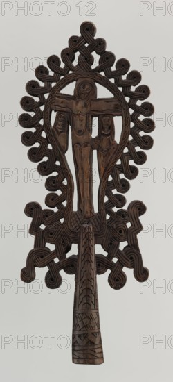 Processional cross, Ethiopian workshop. The pear-shaped frame depicts Christ on the cross with John and Mary standing at the foot in flat relief. Figures are schematically rendered; Christ is bearded, long-haired, with open eyes, head slightly tilted to his right left shoulder, dressed only in a loincloth, feet nailed with a single nail. The pear-shaped frame is decorated with a braided rope pattern and bordered along the outer edge with an openwork braided ornament. The cone-shaped sleeve, flattened on the sides, is decorated with a vertical strip of braid and at the bottom with three horizontal strips of checkered, zigzag, and braided patterns. The arms connecting the sleeve with the cross take the form of an openwork braid. 20th century, Ethiopia. Cross; height 40.5 cm, width 16.0 cm., A carved wooden handheld object with a dark brown color showing a central vertical figure with outstretched arms flanked by two smaller figures, all carved in relief; the top and sides are surrounded ...
